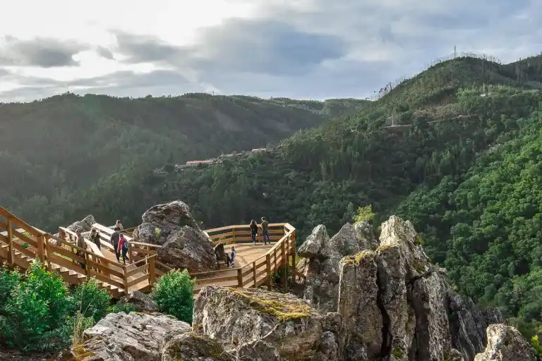 A wooden walkway and stairs descend a cliff, with a view of the surrounding mountains and hills. 
