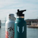 Two Super Sparrow bottles with Lisbon's seaside in the background. One bottle is green, the other white with pink flowers.