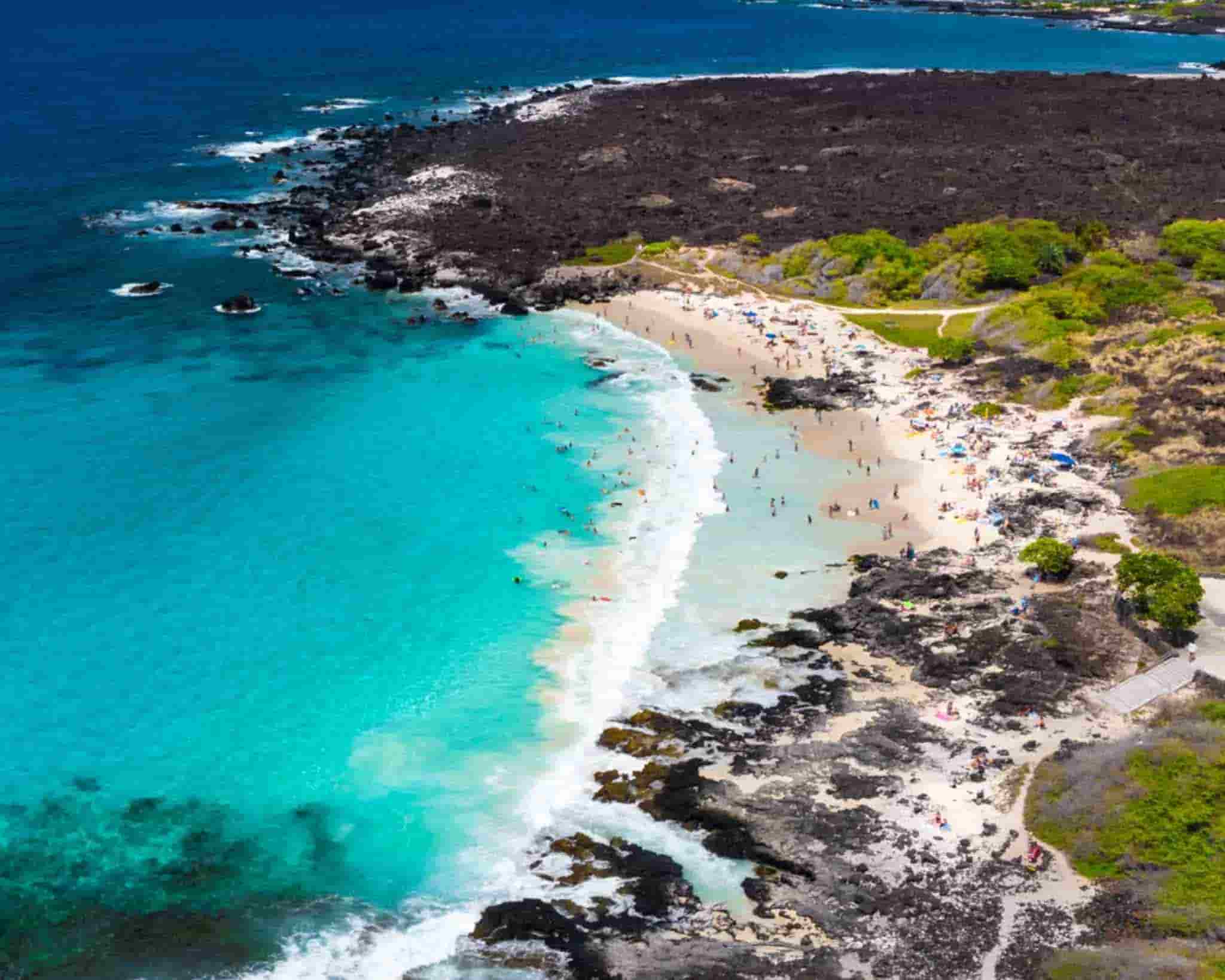 Aerial view of Kua Bay (Manini’owali Beach). The small beach is filled with visitors and surrounded by black rocks.