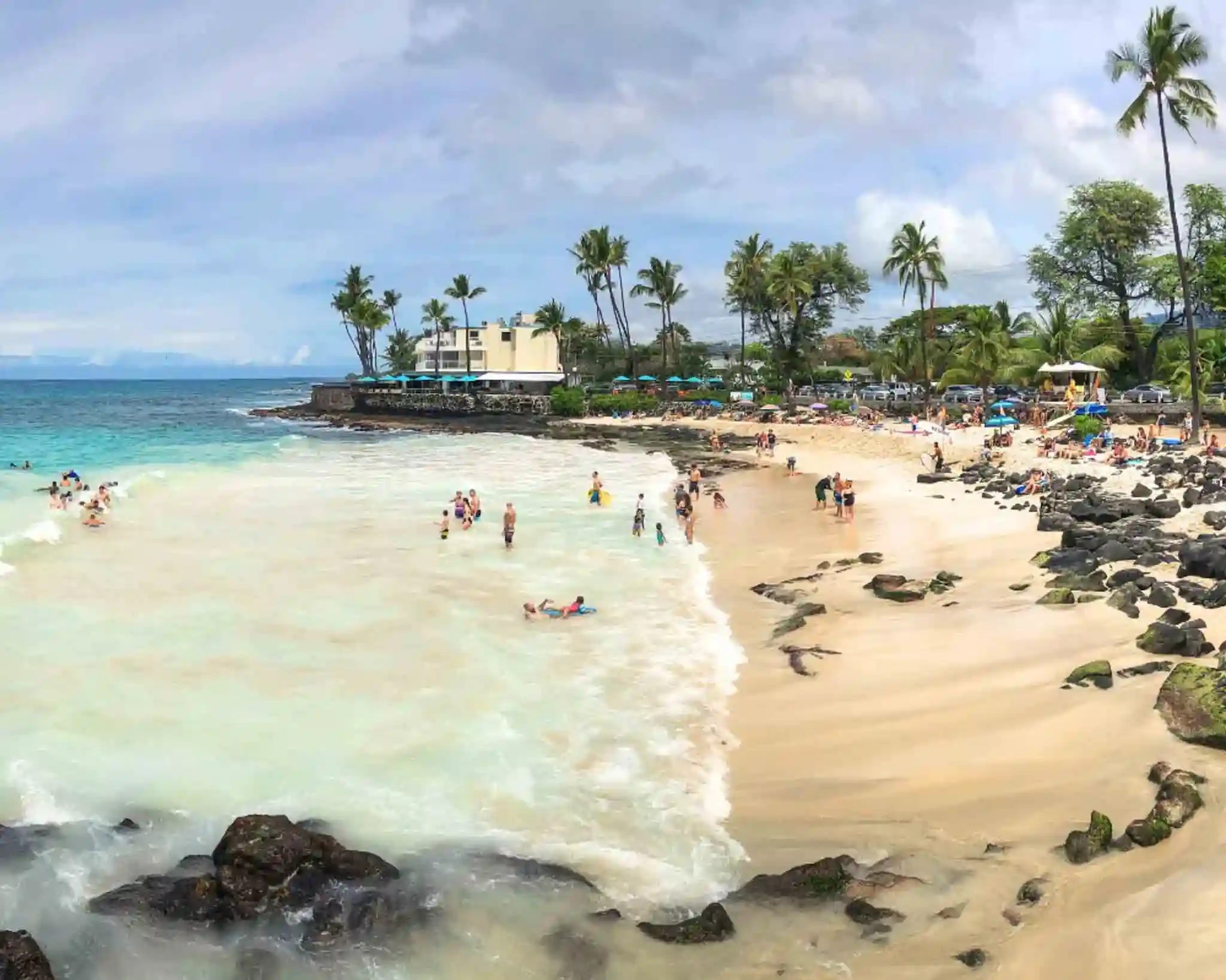 The rocky Magic Sands Beach, surrounded by palm trees and establishments and crowded with visitors.